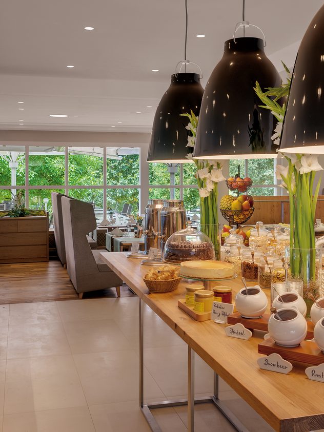 Frühstücksbuffet in einem hellen Restaurant mit Speisen, Getränken, Tischen und großen Fenstern.