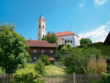 Kirche mit hohem Turm über Garten, Bäumen und Holzhaus in ländlicher Dorfansicht.