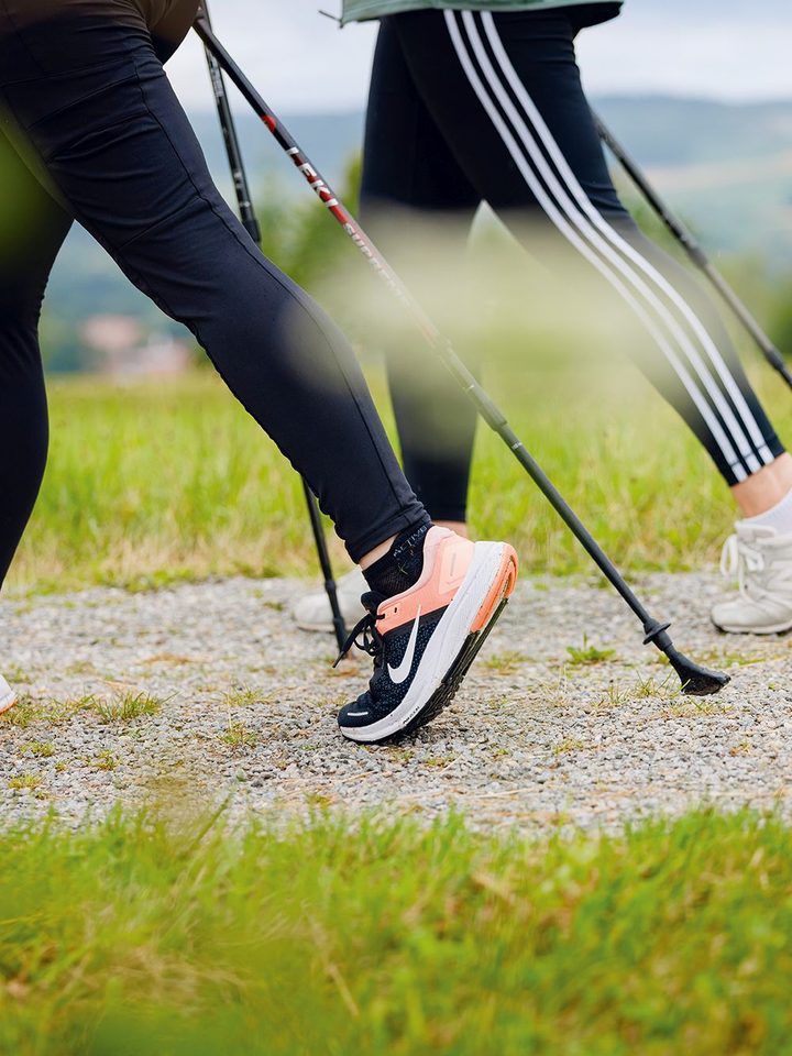 Zwei Personen beim Nordic Walking auf einem Schotterweg in grüner Landschaft.