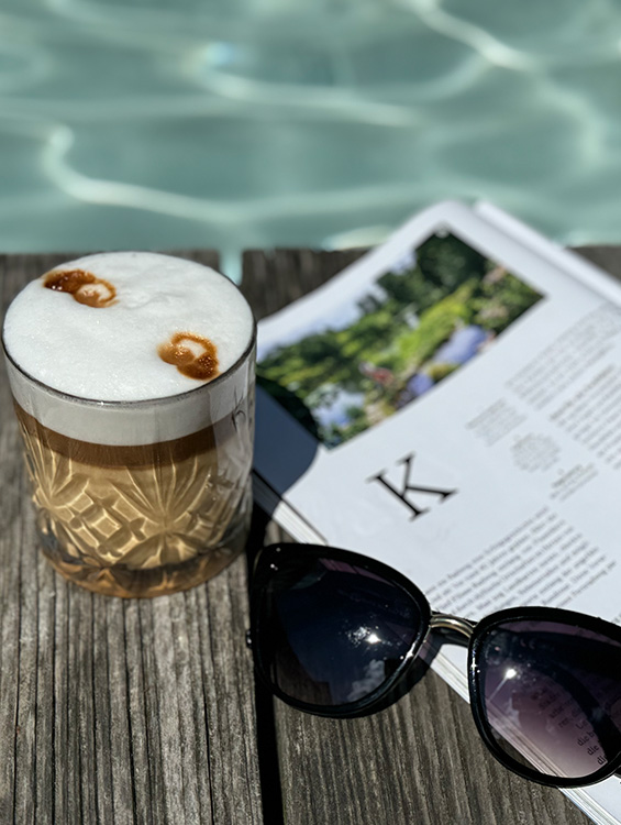 Ein erfrischender Eiskaffee mit Milchschaum und Karamell-Dekoration steht auf einem hölzernen Poolrand. Daneben liegen eine aufgeschlagene Zeitschrift mit einem Gartenfoto und eine schwarze Sonnenbrille. Im Hintergrund schimmert das türkisfarbene Wasser des Pools