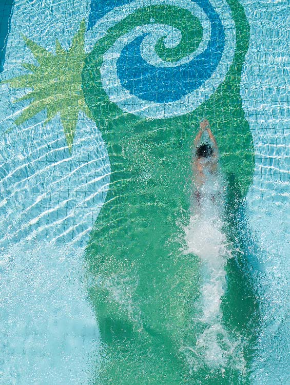 Ein Schwimmer taucht durch das klare Wasser eines Pools, während sich Wellen um ihn herum ausbreiten. Von oben betrachtet, ist ein farbenfrohes Muster auf dem Beckenboden zu sehen, welches das Logo des Sonnenguts zeigt