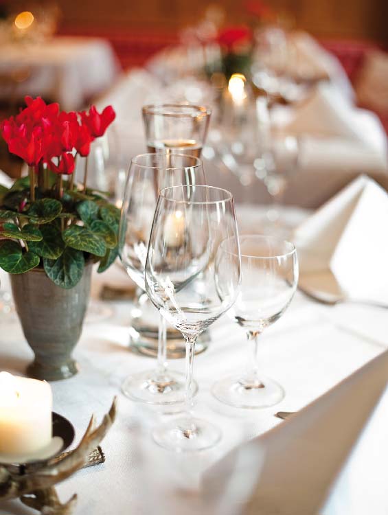 Festlich gedeckter Tisch mit weißen Servietten, edlen Weingläsern und einer stilvollen Tischdekoration aus roten Blumen in einem kleinen Metalltopf. Im Vordergrund brennt eine Kerze, die eine gemütliche Atmosphäre schafft.