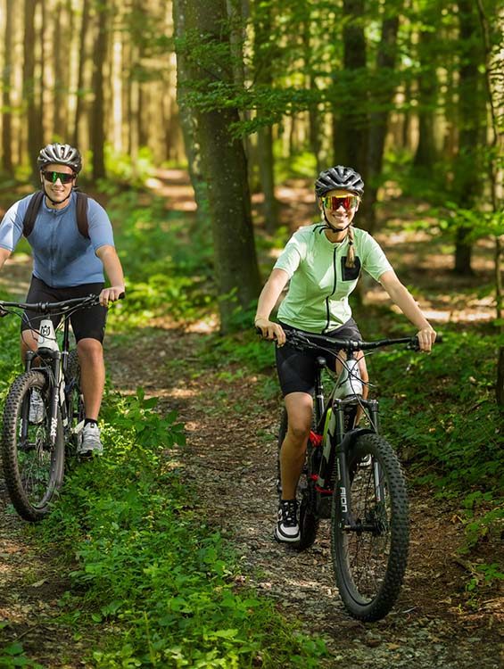 Zwei Personen fahren mit Mountainbikes auf einem schmalen Waldweg. Sie tragen Fahrradhelme und Sportkleidung, während Sonnenlicht durch die Bäume fällt. Die vordere Person trägt eine grüne Radjacke und lächelt, während die hintere Person in einem blauen Shirt folgt.