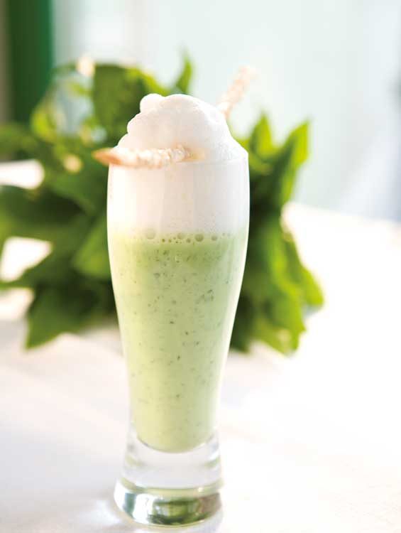Ein hohes Glas mit einem grünen Smoothie oder Shake, getoppt mit einer dicken Schicht weißem Milchschaum. Als Dekoration liegt ein gedrehter Strohhalm oder Trinkröhrchen auf dem Schaum. Im Hintergrund sind unscharfe grüne Blätter zu sehen, was dem Bild eine frische, natürliche Atmosphäre verleiht