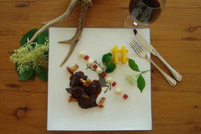 Kunstvoll angerichtetes Wildgericht auf einem weißen Teller mit stilisiertem Hirschmotiv. Das Gericht besteht aus dunklem Fleisch mit Pilzen in Sauce, kleinen Gemüsewürfeln, weißen Cremetupfen, Brokkoliröschen und dekorativ angeordneten Pommes frites. Der Teller ist mit Holzelementen, Blättern, Blüten und einem Hirschgeweih dekoriert. Im Hintergrund liegt Besteck und ein Weinglas auf einem rustikalen Holztisch