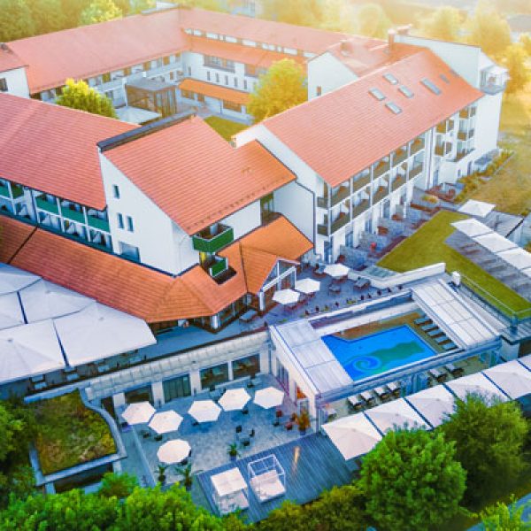 Eine Luftaufnahme des Hotel Sonnengut, eines eleganten Wellnesshotels mit roten Dachziegeln und moderner Architektur. Die Außenanlage umfasst Terrassen mit Sonnenschirmen, ein stilvolles Schwimmbecken und grüne Gärten. Die warme Abendsonne taucht die Szenerie in ein goldenes Licht.