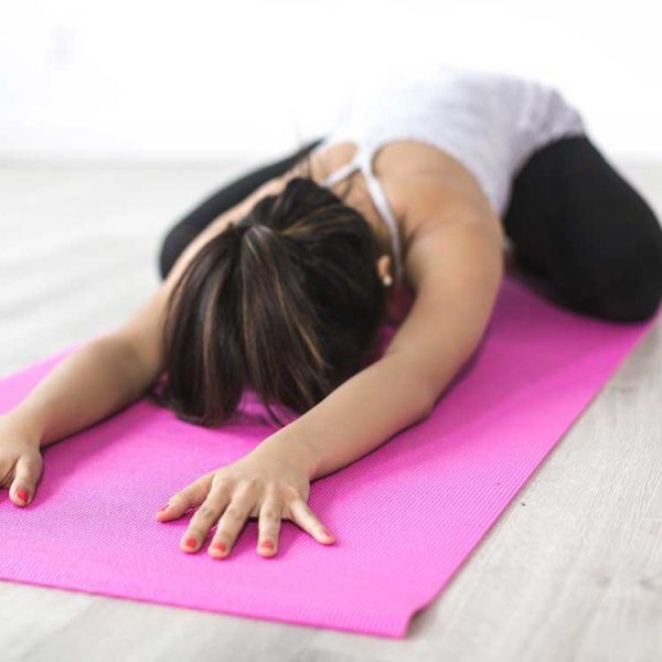 Eine Frau in Sportkleidung auf einer pinken Yogamatte in einem hellen Raum, in der Kindhaltung (Balasana) mit nach vorne ausgestreckten Armen und gesenktem Oberkörper.