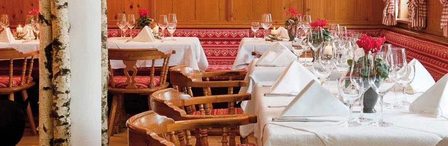 Gemütlich eingerichtete „Hirschstube“ im Wellnesshotel Sonnengut mit liebevoll gedeckten Tischen, Holzvertäfelung, traditionellen Stühlen und roter Alpen-Deko. Weiß eingedeckte Tafeln mit Blumen und Gläsern schaffen eine elegante, warme Atmosphäre.