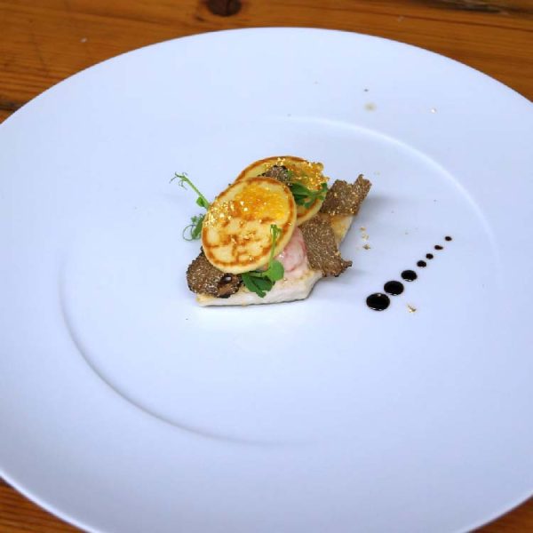 Kunstvoll angerichtete Gourmet-Vorspeise auf einem weißen Teller mit Trüffelscheiben, Mini-Pfannkuchen, Blattgold und feinen Garnierungen; dekorative Balsamico-Tropfen am Tellerran